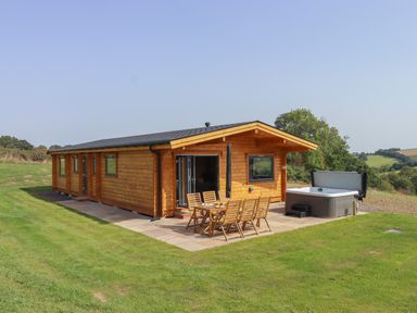 A log cabin with outdoor wooden dining furniture and a hot tub on a patio at Primrose Lodge in Hittisleigh