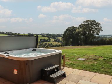 An outdoor hot tub with steps on a tiled patio overlooking green fields and trees at Primrose Lodge in Hittisleigh