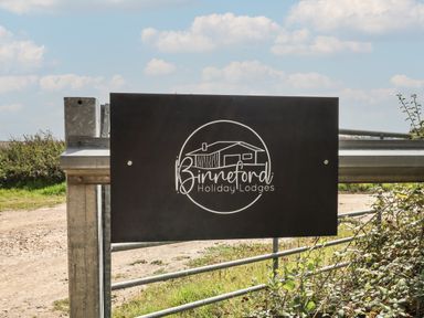 A metal gate with a black sign showing a house and the text Binneford Holiday Lodges in a rural outdoor setting