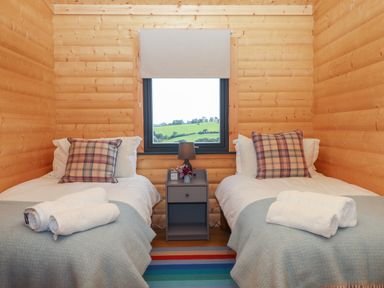 A wooden bedroom with two single beds a gray nightstand a window showing green fields and a striped rug at Primrose Lodge in Hittisleigh
