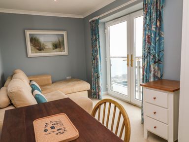 A living room with a beige sofa a wooden dining table with a chair a small white cabinet and glass doors with floral curtains at Looe Island View in Downderry