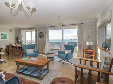 A living room with blue chairs a wooden coffee table and a dining area with a sea view at Looe Island View in Downderry