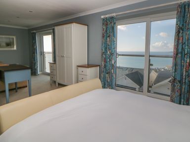 A bedroom with a bed wardrobe and drawers next to sliding glass doors with blue floral curtains overlooking the sea at Looe Island View in Downderry
