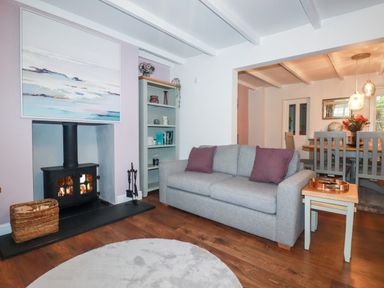 A living room with a grey sofa and purple pillows next to a fireplace and a bookshelf with a dining area visible in the background at Still Waters in Pentewan