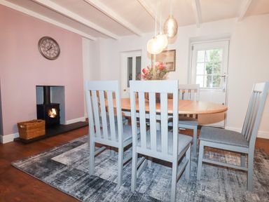 A dining room with a wooden table and six gray chairs next to a wood burning stove and wall clock at Still Waters in Pentewan