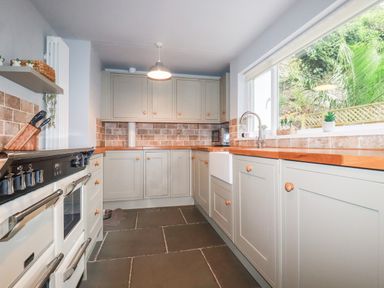 A kitchen with gray cabinets wooden countertops a stove a sink and a large window showing greenery outside at Still Waters in Pentewan
