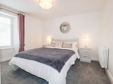 A bedroom with a bed with gray blanket and pillows bedside tables with lamps a round wall mirror window with curtains and a radiator at Still Waters in Pentewan
