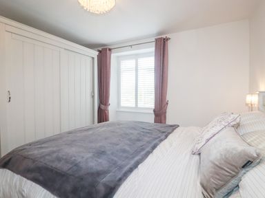 A bedroom with a bed covered in a grey blanket and pillows next to a window with purple curtains and white wardrobe at Still Waters in Pentewan