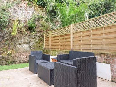 An outdoor patio with black wicker chairs and a table next to a wooden fence and stone wall at Still Waters in Pentewan