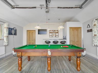 A game room with a pool table in the center three bar stools at a home bar a dartboard on the wall and two wooden doors at Rose Cottage in Muff County Donegal