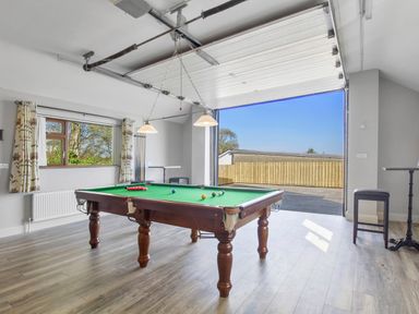 A room with a pool table under hanging lights a window with floral curtains an open garage door and a tall stool at Rose Cottage in Muff County Donegal