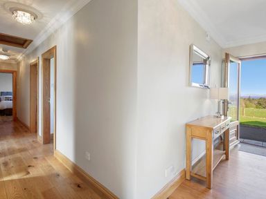 A hallway with wooden floors and doors leading to rooms and a wooden table with a lamp next to an open front door showing a grassy outside area at Rose Cottage in Muff County Donegal