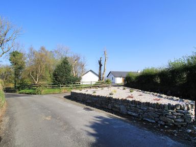A rural road with a stone wall garden bed and white buildings in the background at Rose Cottage in Muff County Donegal