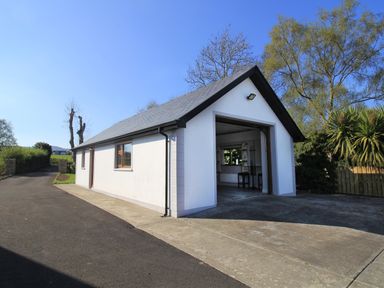 A white single-story outbuilding with an open garage door next to a paved driveway and surrounded by trees at Rose Cottage in Muff County Donegal