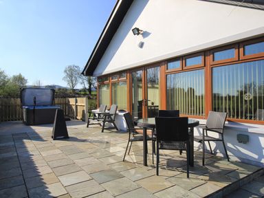 An outdoor patio with a round glass table and four black chairs a hot tub and two wooden chairs next to a white building with large windows at Rose Cottage in Muff County Donegal