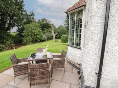 An outdoor patio with a glass table and four wicker chairs next to a white textured building surrounded by grass and trees at Grove Cottage in Leominster