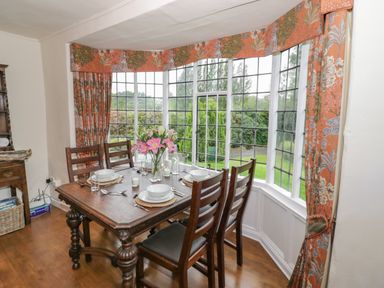 A dining area with a wooden table and chairs set for four near bay windows with floral curtains at Grove Cottage in Leominster
