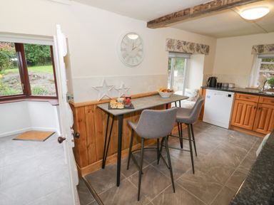 A kitchen with a high table and two stools a clock on the wall and a window seat at Grove Cottage in Leominster