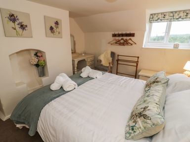 A bedroom with a bed made with white linens and floral pillows a chair a vanity table and window at Grove Cottage in Leominster