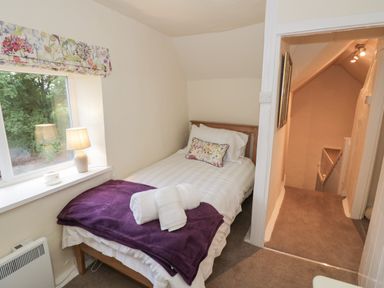 A small bedroom with a single bed a purple blanket and towels next to a window with a floral blind at Grove Cottage in Leominster