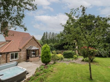 A garden with trees and bushes next to a stone house with a tiled roof and a hot tub on the patio at Grove Cottage in Leominster