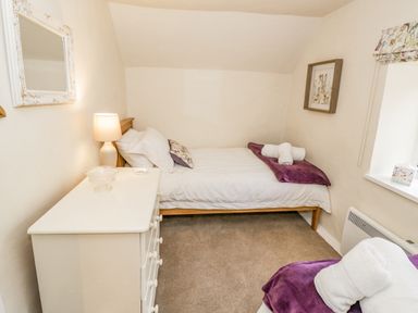 A bedroom with a bed with white linens and purple blanket next to a white dresser with lamp at Grove Cottage in Leominster