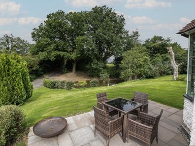 A patio with a glass table and four wicker chairs overlooking a grassy yard and trees at Grove Cottage in Leominster
