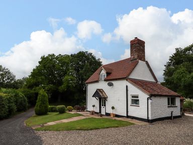 A white cottage with a red tiled roof and a chimney surrounded by grass and trees at Grove Cottage in Leominster