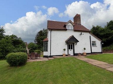 A white cottage with a red roof and chimney on a grassy lawn with a paved pathway and outdoor seating at Grove Cottage in Leominster