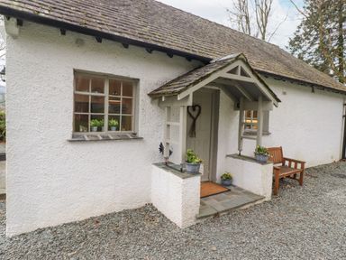 An entrance door with a small porch and potted plants next to windows and a wooden bench outside a white textured cottage