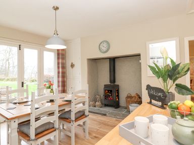 A dining area with a wooden table and white chairs near a wood burning stove and a kitchen counter with a tray of cups and a fruit basket at Larch Cottage at Esthwaite Water in Hawkshead