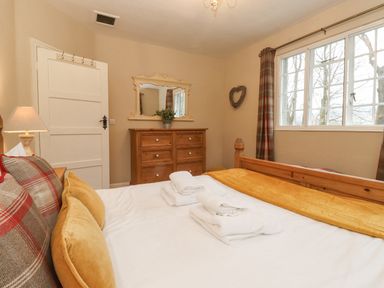 A bedroom with a wooden bed yellow pillows white towels a wooden dresser with a mirror and a window with plaid curtains at Larch Cottage at Esthwaite Water Hawkshead