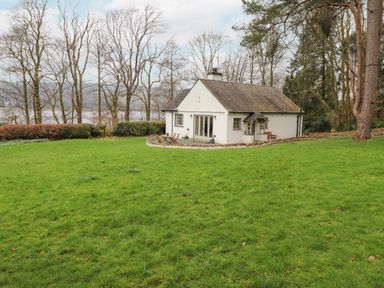 A white cottage with outdoor chairs and tables surrounded by grass and trees near a lake at Larch Cottage at Esthwaite Water Hawkshead