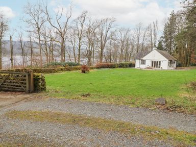 A house with a gravel driveway and fenced yard near trees and a body of water at Larch Cottage at Esthwaite Water in Hawkshead