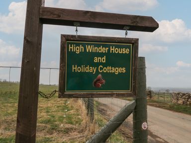 A wooden sign for High Winder House and Holiday Cottages on a post beside a rural road with fields and a wire fence