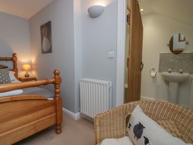 A bedroom with a wooden bed frame a bedside table with lamp a wicker chair with a cushion and an open door leading to a bathroom with a sink and mirror at Winder Barn in Askham Cumbria