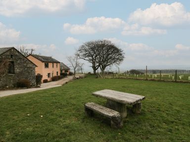 A grassy field with a wooden picnic table and benches near stone and peach buildings with trees in the background at Winder Barn in Askham Cumbria