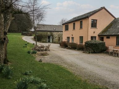 A courtyard with wooden chairs and table near a two-story peach colored house and a stone building at Winder Barn in Askham Cumbria