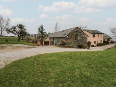 A gravel path leading to stone and peach-colored buildings with grassy fields and trees in the background at Winder Barn in Askham Cumbria