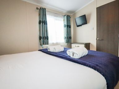 A bedroom with a bed with rolled towels a window with curtains a wall mounted television a wooden door and a small drawer unit at Lake View 10 in Par
