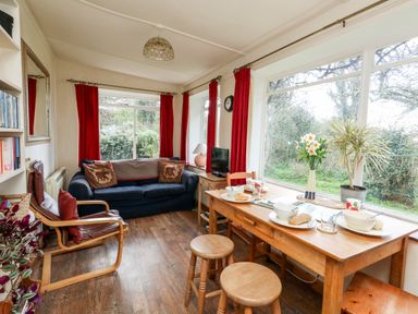 A living room with a blue sofa red curtains a wooden dining table and large windows overlooking greenery at Eweleaze in West Bexington