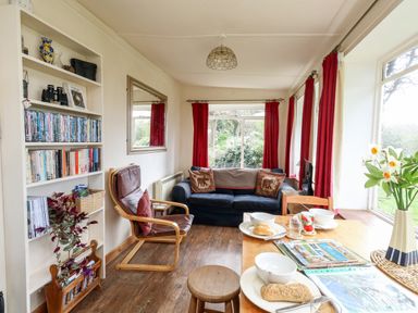A living room with a blue sofa red curtains a wooden chair table set with food and books on a shelf at Eweleaze in West Bexington
