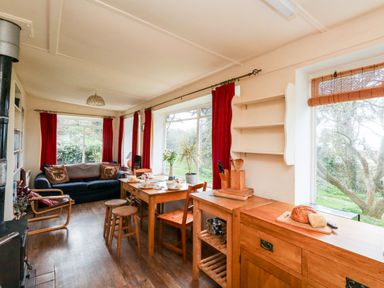 A dining area with a wooden table and stools next to large windows with red curtains a blue sofa and wooden armchair near a black stove in a living room at Eweleaze West Bexington