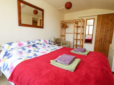 A bedroom with a bed covered with a red blanket and colorful sheets two towels on the bed a wooden shelving unit a wicker chair a mirror on the wall and a wooden door at Eweleaze in West Bexington