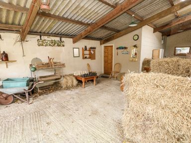 A rustic room with hay bales a wooden table with a chess set a wheelbarrow and wooden chairs at Eweleaze in West Bexington