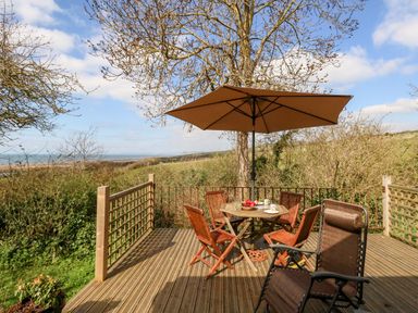 A wooden deck with a round table and four chairs under a large umbrella and a reclining chair overlooking greenery and a distant sea view at Eweleaze in West Bexington