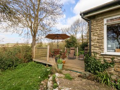 A wooden deck with chairs a table and umbrella next to a stone house surrounded by trees and plants at Eweleaze in West Bexington