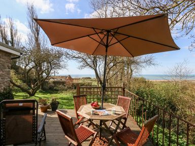 An outdoor wooden deck with a table and chairs under a large umbrella overlooking a garden and the sea at Eweleaze West Bexington
