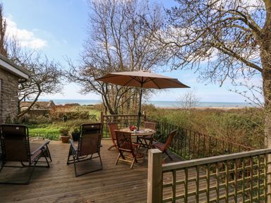 A wooden deck with chairs a table and an umbrella overlooking trees and the sea at Eweleaze in West Bexington