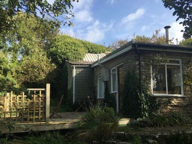 An exterior view of a stone cottage with a wooden deck and garden chairs surrounded by trees and bushes at Eweleaze in West Bexington